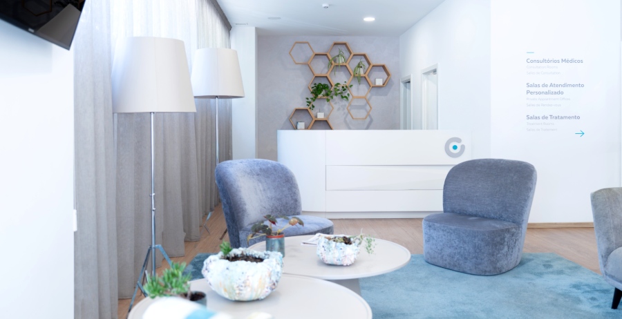 Heller, freundlich gestalteter Wartebereich der Ferticentro Kinderwunschklinik in Coimbra mit grauen Sesseln, runden Couchtischen, bodentiefen Vorhängen und weißem Empfangstresen mit Wandschmuck