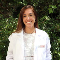 Dr. Marina Diaz von der Clínica Tambre im weißen Laborkittel vor grünen Büschen.