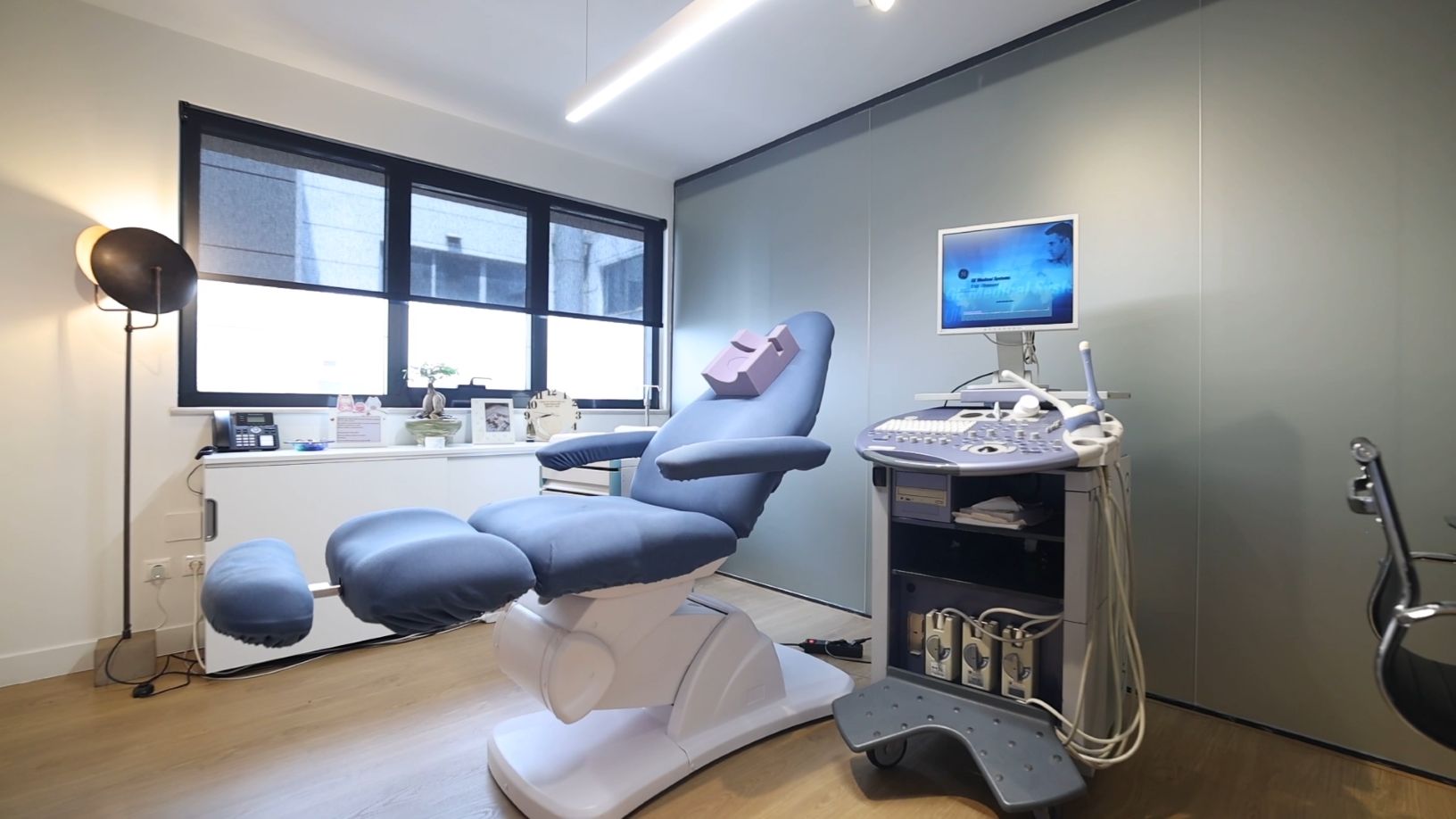 Untersuchungs- und Ultraschallraum in einer modernen Kinderwunschklinik mit einem gynäkologischen Stuhl, einem Ultraschallgerät und einem großen Fenster für Tageslicht.