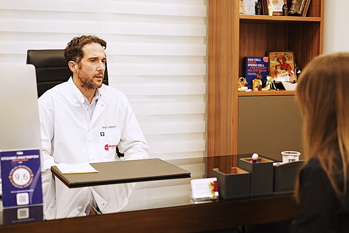 Dr. Panagiotis Drakopoulos berät Patientin in Reproduktionsmedizin: Arzt in weißem Kittel am Schreibtisch, Frau mit langen Haaren, Bücherregal im Hintergrund.