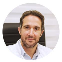 Profilfoto von Dr. Panagiotis Drakopoulos, IVF-Spezialist: Mann mit grauem Bart in weißem Kittel, sitzend vor schwarzem Stuhl