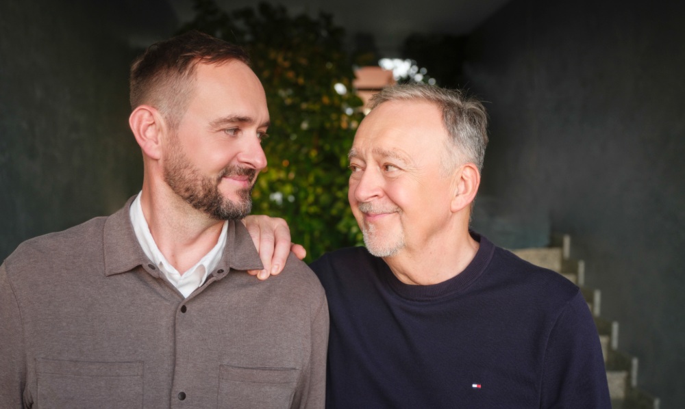 Dr. Milan Mrázek (älterer Mann im blauen Pullover) und sein Sohn Martin Mrázek (jüngerer Mann im braunen Hemd), Gründer der GYNEM-Fruchtbarkeitsklinik, lächeln gemeinsam vor grünem Hintergrund.